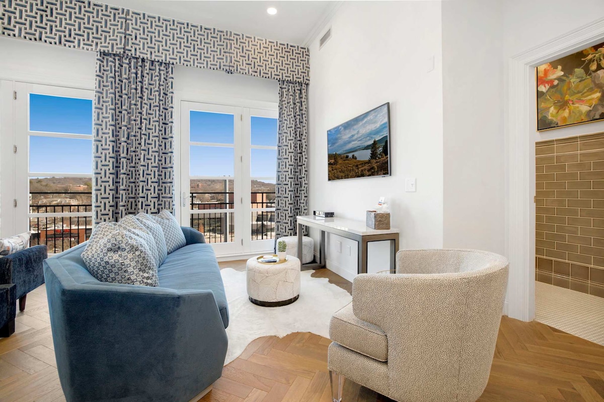 A cozy living area is furnished with a blue sectional sofa, a circular coffee table, and a light grey accent chair. Large windows adorned with patterned curtains offer abundant natural light and a view of the surroundings. A wall-mounted television is positioned for easy viewing.
