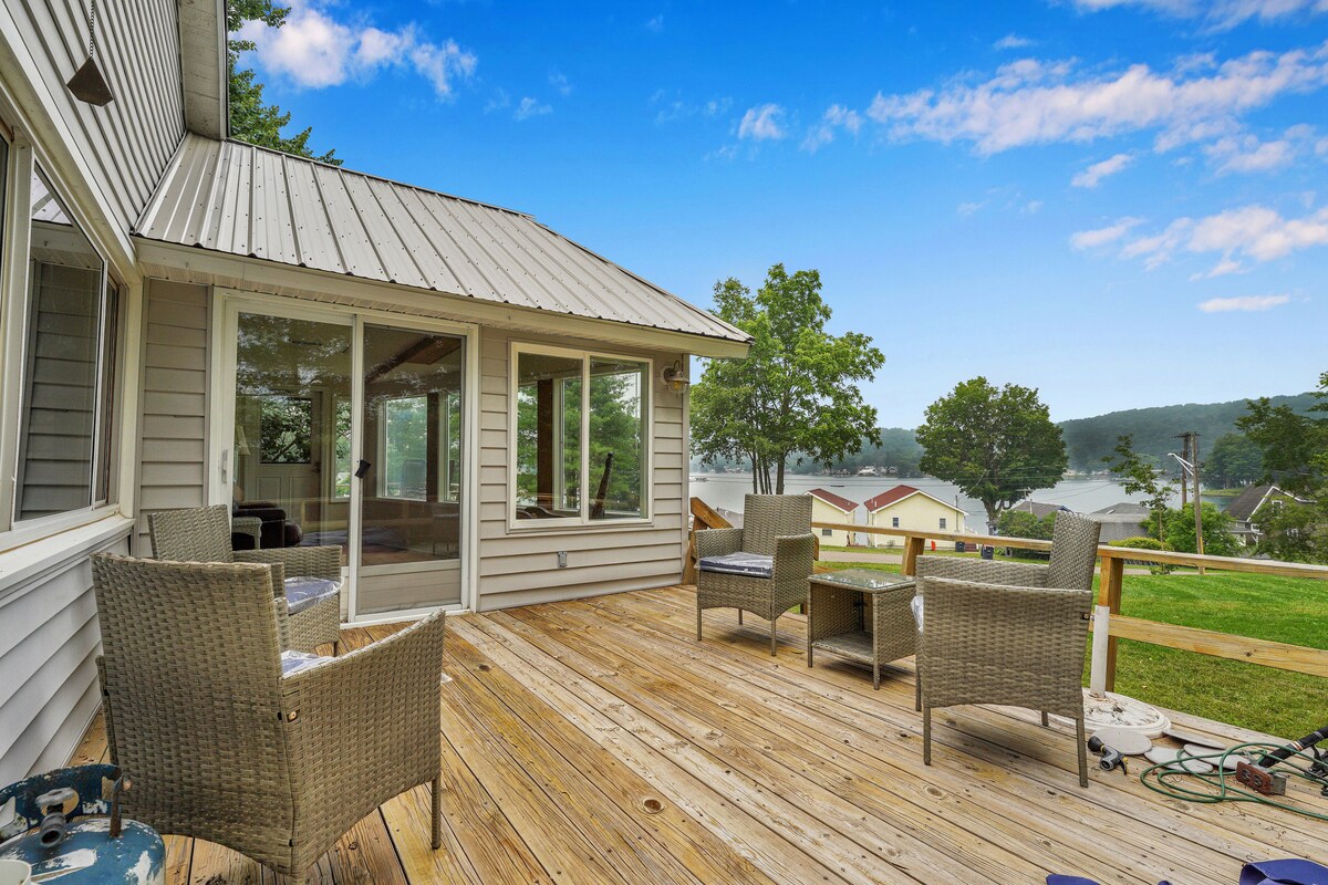 A spacious deck features multiple wicker chairs arranged for relaxation, overlooking scenic lake views. Large glass windows and sliding doors provide natural light and connect the indoor sunroom to the outdoor space. The surrounding greenery enhances the serene atmosphere of the lakeside home.