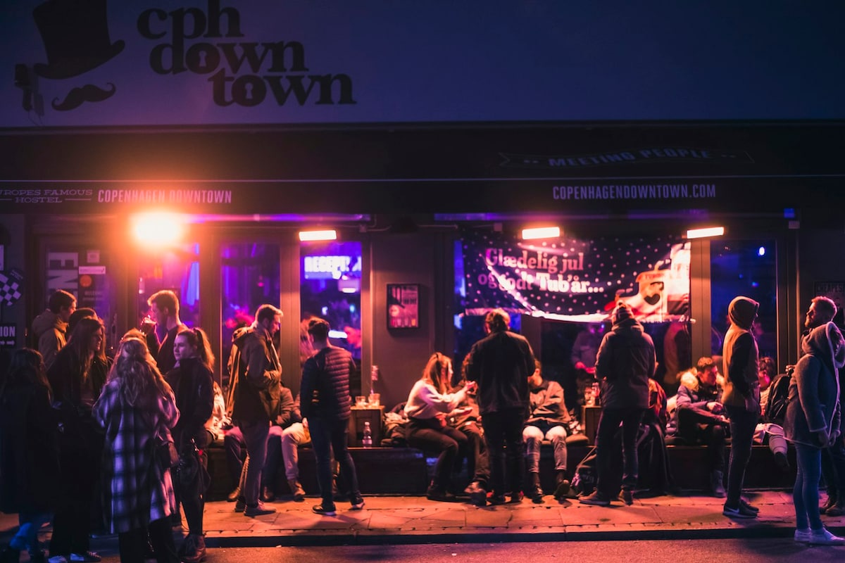 The exterior of a lively bar is shown, with vibrant neon lights casting a colorful glow. Groups of people are seated and standing outside, engaged in conversation. The atmosphere appears social, with a mix of patrons enjoying the night.