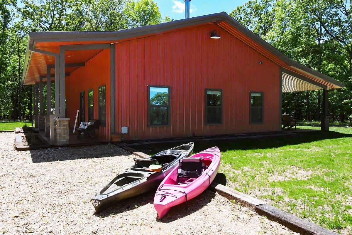 A warm-colored cabin is shown surrounded by greenery. Three kayaks, two in muted tones and one in pink, rest on a gravel area in front of the cabin, with large windows reflecting the natural landscape.