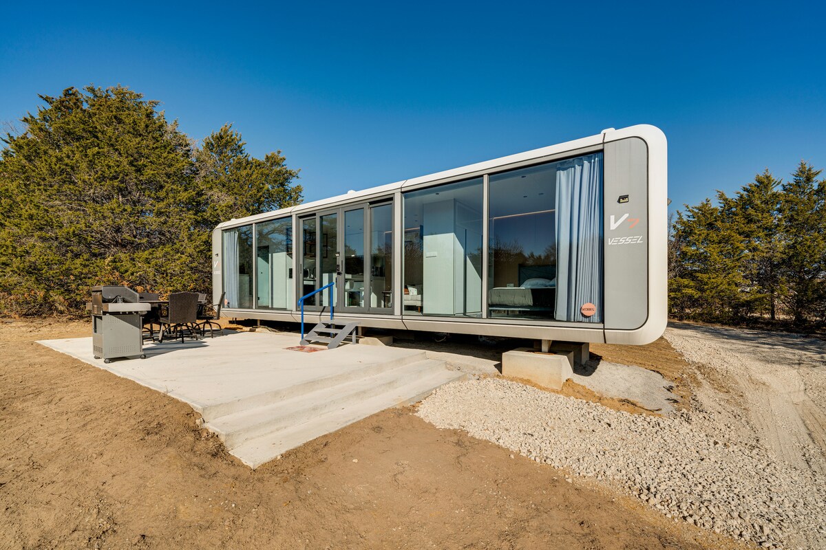 The studio's exterior is characterized by a modern design, featuring a long glass wall that offers expansive views of the surrounding tree line. A patio area is visible, equipped with a gas grill and outdoor seating, creating an inviting space for relaxation.