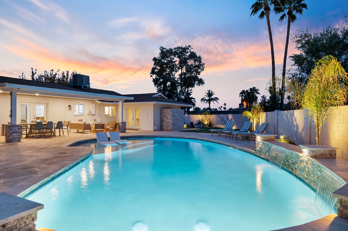 A tranquil backyard scene showcases a sparkling pool, surrounded by comfortable lounge chairs and a shaded patio area. Gentle lighting highlights the water's surface as the sky transitions to dusk with soft hues. Palm trees frame the setting, contributing to a relaxing outdoor atmosphere.
