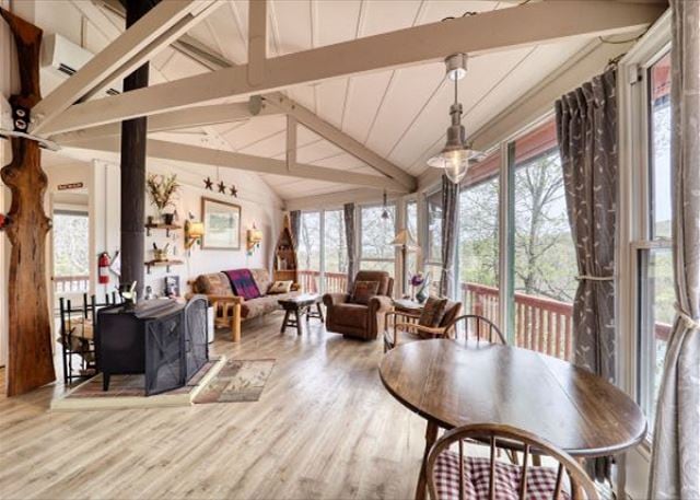 An open living area features large windows that allow for natural light and views of the surrounding landscape. A wood-burning stove is placed near a seating arrangement, while a dining table sits centrally among rustic decor and comfortable furnishings.