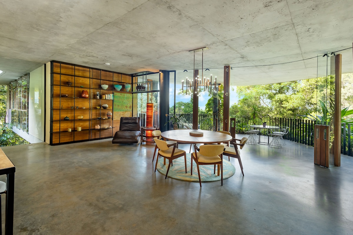 An open-plan dining area features a round wooden table surrounded by four chairs. A large shelving unit displays colorful decorative items. Floor-to-ceiling glass doors provide views of the lush surroundings, enhancing the connection to the rainforest.