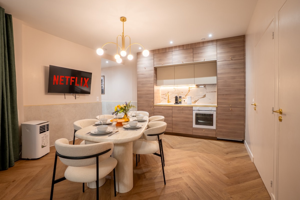 A modern dining area is presented, featuring a round table surrounded by cream-colored chairs. A stylish light fixture hangs overhead, while a television displaying the Netflix logo is mounted on the wall. Kitchen cabinets with a neutral finish provide a clean, contemporary look.