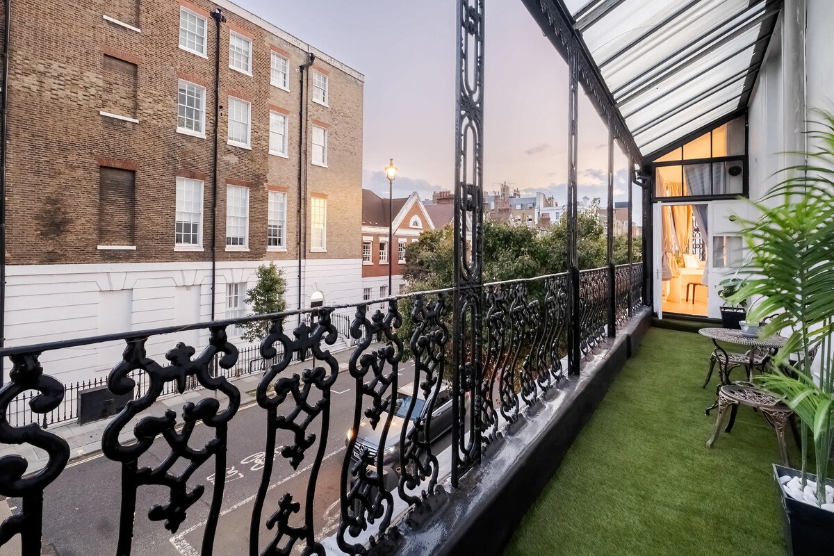 A chic private balcony features intricate wrought iron railings and lush artificial grass. Two small chairs accompany a table, offering a serene view of the surrounding street and buildings. Soft evening light casts a warm glow, enhancing the inviting outdoor space.