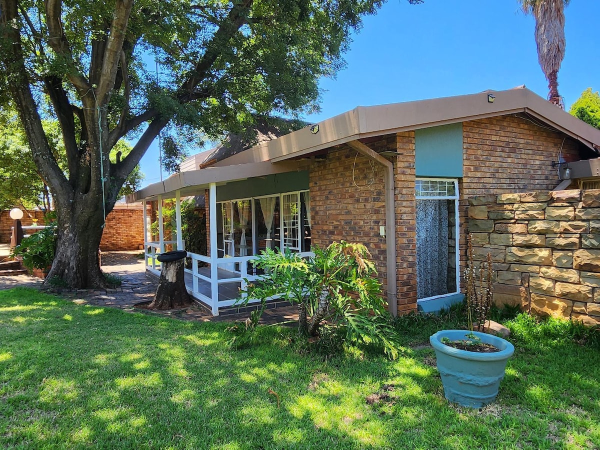 The exterior of the unit is shown, featuring a brick facade and covered porch area. Lush greenery surrounds the building, with large trees offering shade. A stone wall adds texture to the landscape, and a potted plant enhances the outdoor space.