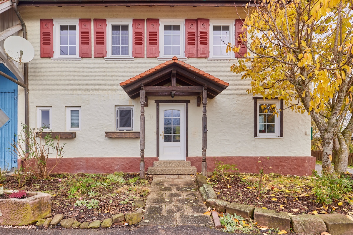 A charming entrance is framed by a wooden canopy, featuring a central door with a decorative light fixture. Windows are adorned with red shutters, with a landscaped area including flower beds and a pathway leading to the front. A basketball hoop is visible on one side.
