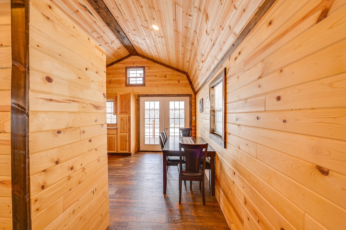 A warm and inviting interior showcases natural wood paneling and a high ceiling. The dining area features a long table surrounded by several chairs, with double doors leading outside. Natural light filters in through windows, enhancing the spacious feel of the cabin.