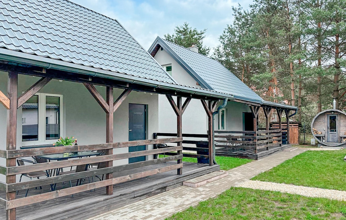 A two-story holiday home is depicted, featuring a gray exterior with a sloped roof. A spacious wooden veranda encompasses the front, furnished with a table and chairs. The surrounding area includes a neatly paved walkway and greenery, enhancing the welcoming environment.