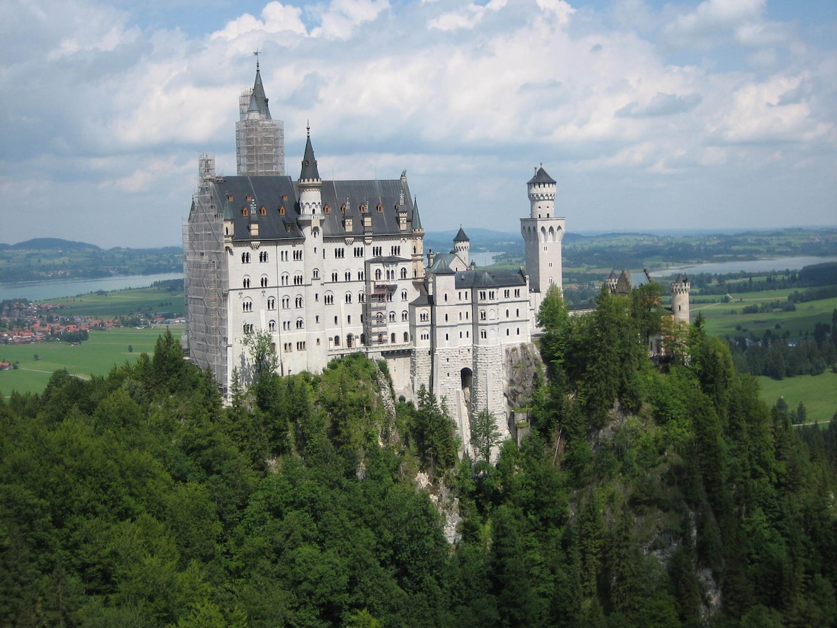 A grand castle, with intricate architectural features, is perched on a hillside surrounded by lush greenery. The tall spires and towers rise against a backdrop of a bright blue sky dotted with clouds. Scenic valleys and fields stretch out in the distance, enhancing the view.