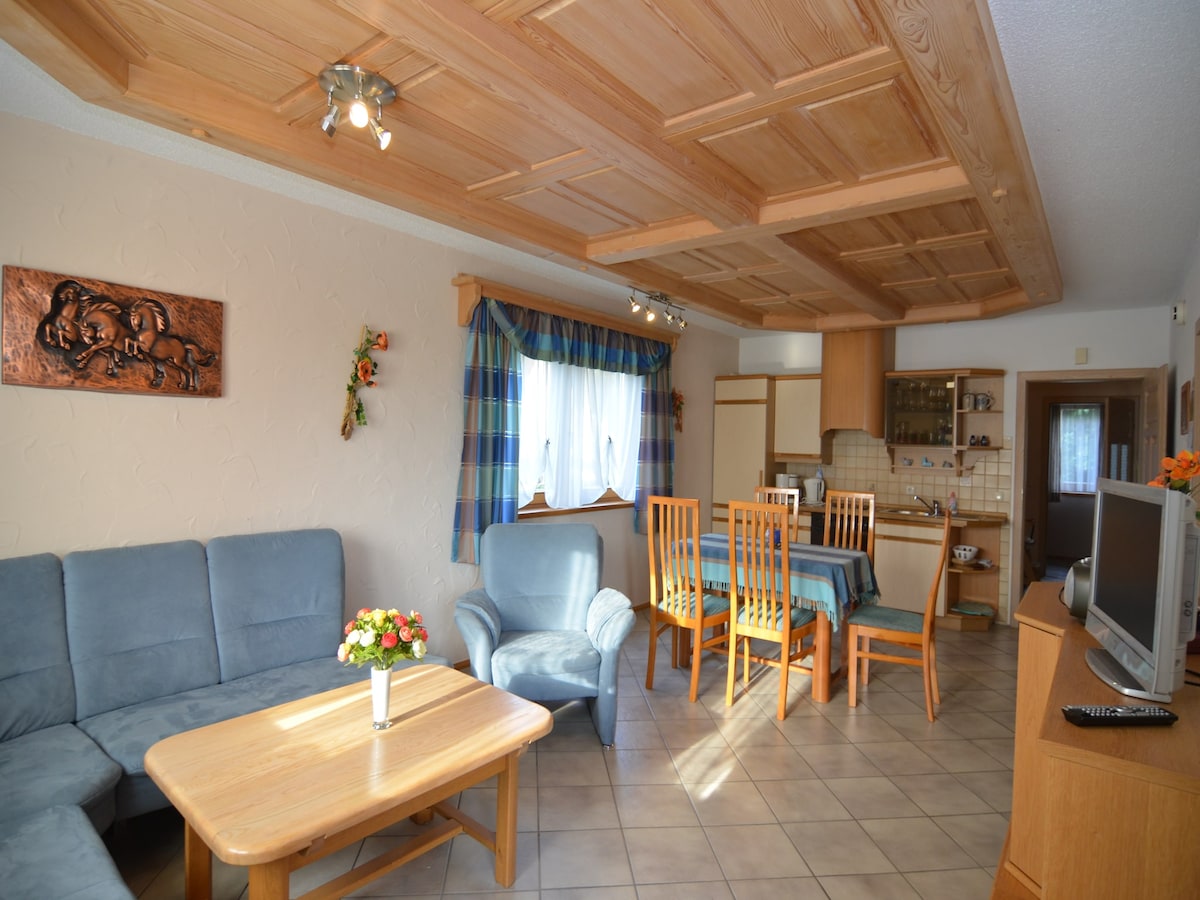 An open living and dining area features a blue sofa set, a light wooden coffee table, and a dining table surrounded by six chairs. A window draped in patterned fabric allows natural light, complementing the wooden ceiling beams and wall decorations.