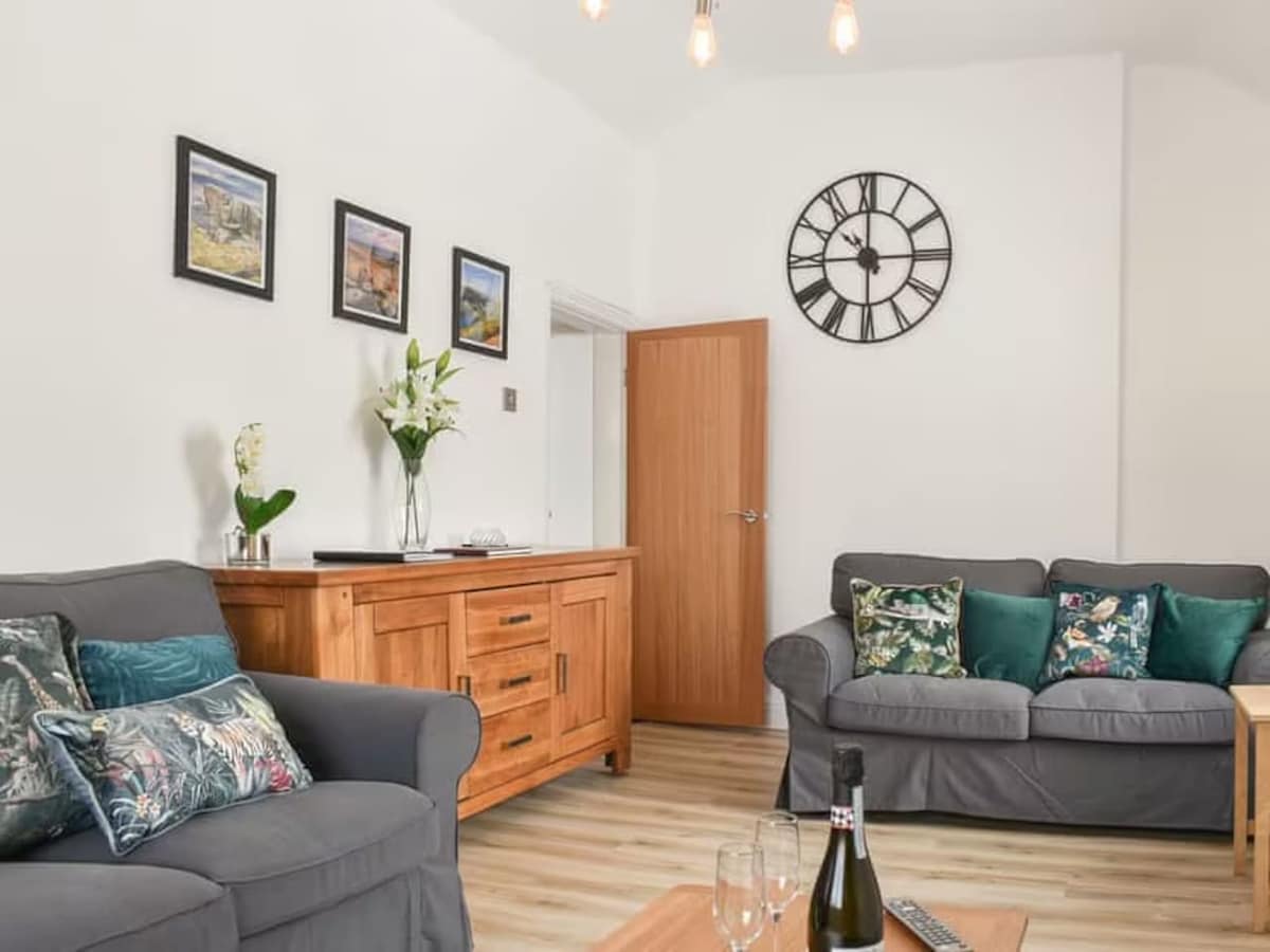 The living room features two large sofas adorned with decorative cushions. A wooden sideboard holds a vase with flowers. Framed artwork decorates the walls, and a wall clock adds a modern touch. Natural light enhances the warm wooden flooring.