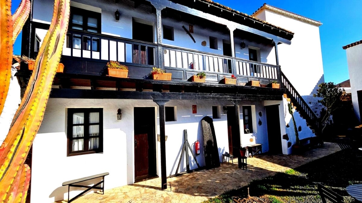 The exterior of Casa Rural Isaitas displays a traditional two-story structure with whitewashed walls and dark wooden balconies. Planters with blooming flowers adorn the balcony, while a stone pathway leads to various entrances. A staircase rises alongside the building, enhancing accessibility.