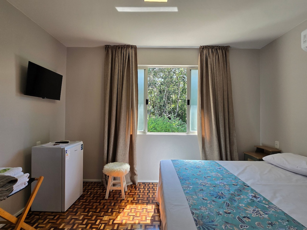 A well-lit room features large windows with greenery visible outside, enhancing natural light. The space includes a cozy bed dressed in white linens and a patterned throw. A compact refrigerator and a TV are also present, along with a small stool and neatly arranged towels.