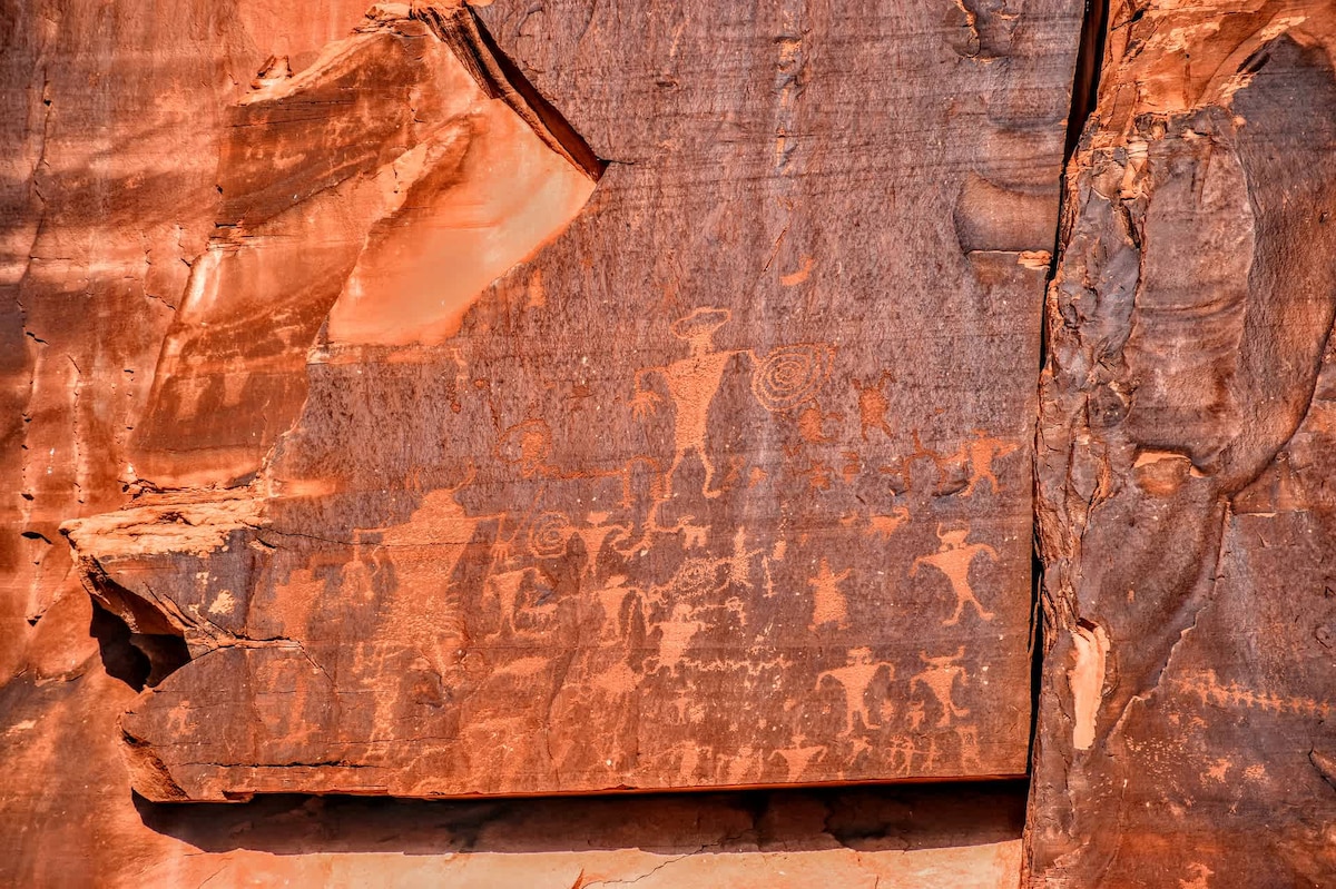 Ancient petroglyphs are etched into a large rock face, showcasing various figures and symbols that reflect the region's historical significance. The reddish-brown sandstone surface exhibits a natural texture, with faint traces of artistry against the rugged landscape.