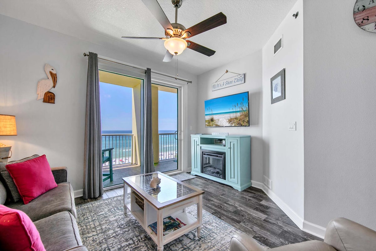 A living area features a comfortable gray sofa with decorative pillows, a coffee table with a light wood finish, and a television mounted on a pastel blue entertainment center. Soft light enters through sliding glass doors, leading to a balcony with views of the Gulf.