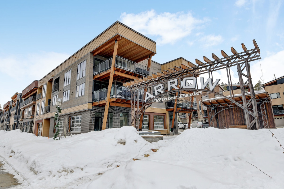 A contemporary multi-story townhome complex is showcased, featuring a prominent sign that reads 'ARROW' at the entrance. Snow blankets the ground, and balconies line the upper floors, with wooden accents enhancing the modern design. Clear blue skies provide a serene backdrop.