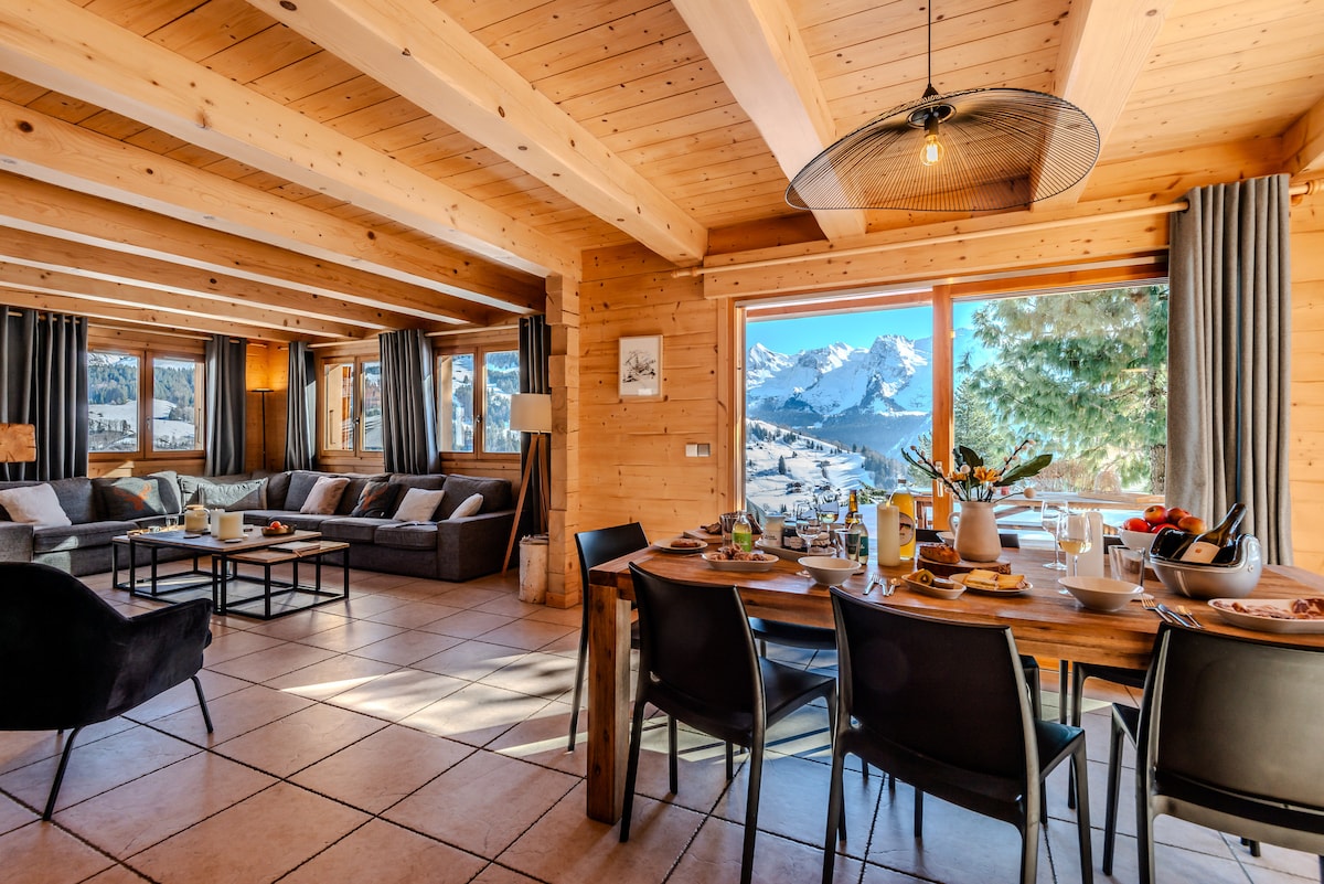 The spacious living area features wooden beams and large windows offering panoramic views of the Aravis mountains. A comfortable seating arrangement includes a sofa and armchairs, while a dining table is set for a meal. Natural light fills the room, enhancing the warm ambiance.