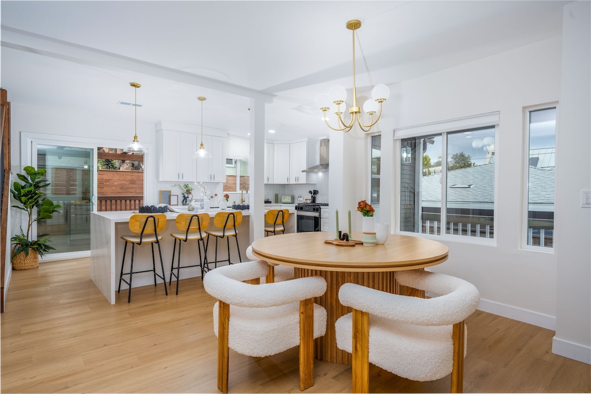 A modern dining area features a round wooden table surrounded by plush white chairs. Bright windows provide natural light, while a stylish chandelier hangs above. The open kitchen is visible in the background, showcasing sleek cabinetry and bar seating.