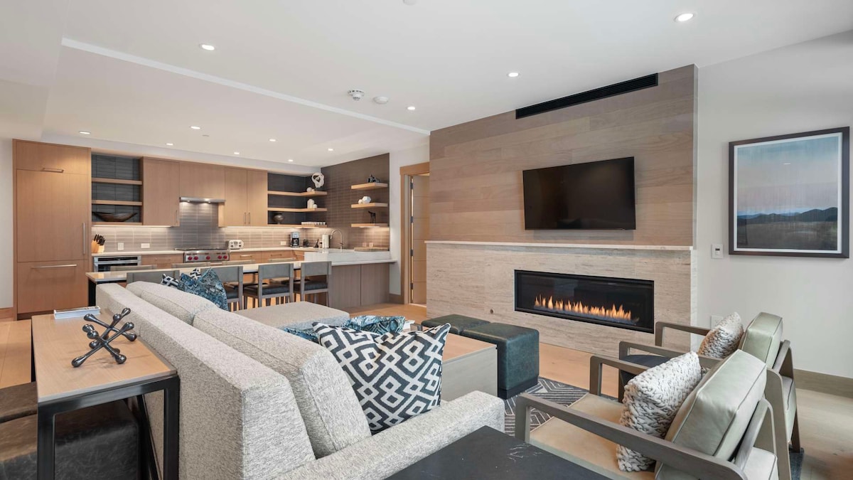 A modern living area features a sectional sofa paired with lounge chairs, emphasizing comfort and space. A sleek gas fireplace is mounted on the wall, complemented by a flat-screen TV. The open layout leads to a kitchen with contemporary cabinetry and appliances.