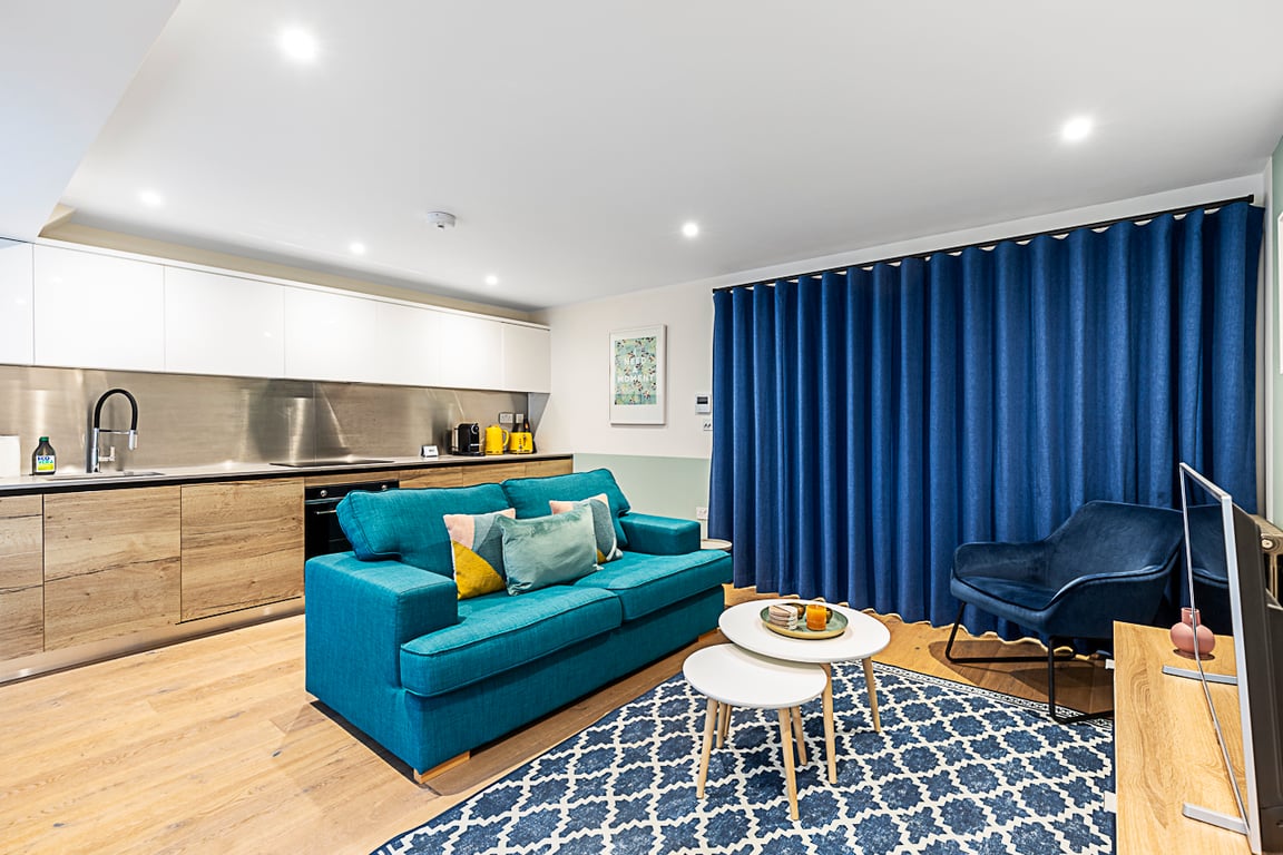 A modern living space features a deep blue sofa complemented by colorful cushions. A light wooden coffee table sits on a patterned blue and white rug. In the background, a sleek kitchen with white cabinetry and stainless steel finishes is visible, along with a plush armchair and dark blue curtains.