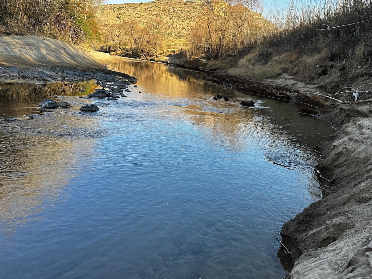 The calm waters of a river are reflected in the gentle light, creating a serene atmosphere. The riverbanks are framed by trees, with rocky outcrops visible along the water's edge. A backdrop of rolling hills is partially covered with sparse vegetation.