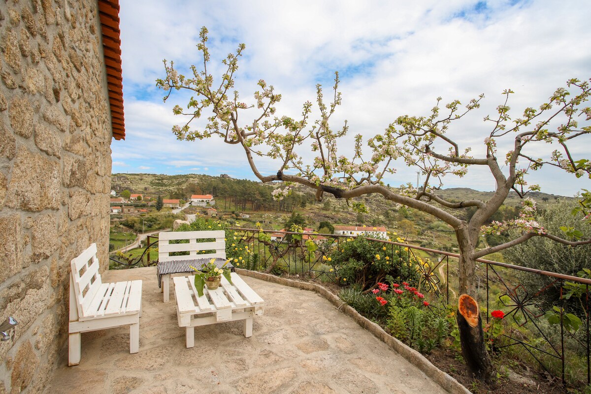 A charming terrace area features two white wooden chairs with a small table, surrounded by a stone surface. A flowering tree extends over the space, enhancing the view of the countryside, which includes fields and distant buildings under a partially cloudy sky.