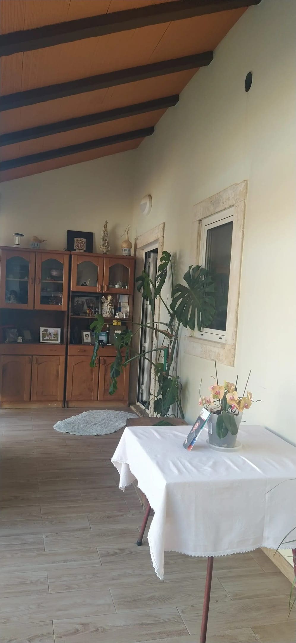 A sunlit entryway features a wooden cabinet displaying decorative items and plants. A small table with a white tablecloth and a vase of flowers is positioned nearby. The space is enhanced by high, exposed beam ceilings and a window that allows natural light to fill the area.