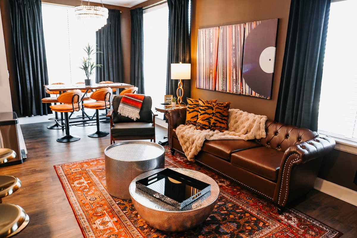 A stylish living area features a plush brown leather sofa adorned with a textured throw. A chic round coffee table sits on a decorative area rug. Large windows draped with dark curtains allow natural light to filter in, while modern artwork adds a vibrant touch to the wall.