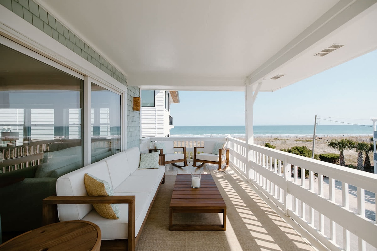 A covered porch is presented with comfortable outdoor seating arrangements, including a white sofa and wooden chairs. A coffee table sits at the center. The ocean is visible in the background, offering a serene seaside view framed by the porch railing.