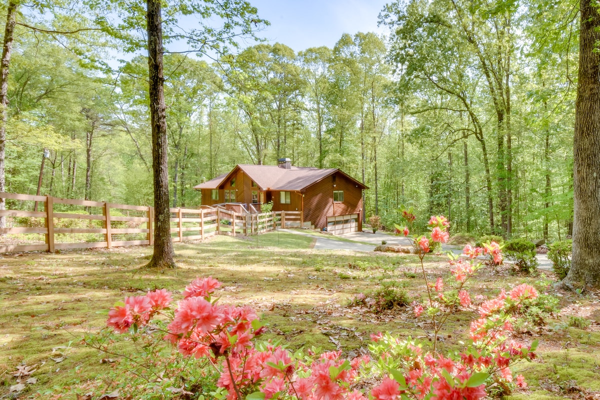 A rustic cabin is surrounded by lush greenery and tall trees. Vibrant pink azaleas provide a pop of color in the foreground, contrasting with the earthy tones of the wooden structure. A spacious yard and a fence enhance the sense of privacy in this natural setting.