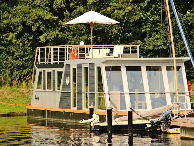 A modern houseboat is anchored at the water's edge, featuring large windows that allow abundant natural light. A rooftop terrace with a sunshade provides outdoor seating. Natural greenery surrounds the vessel, enhancing its serene environment by the river.