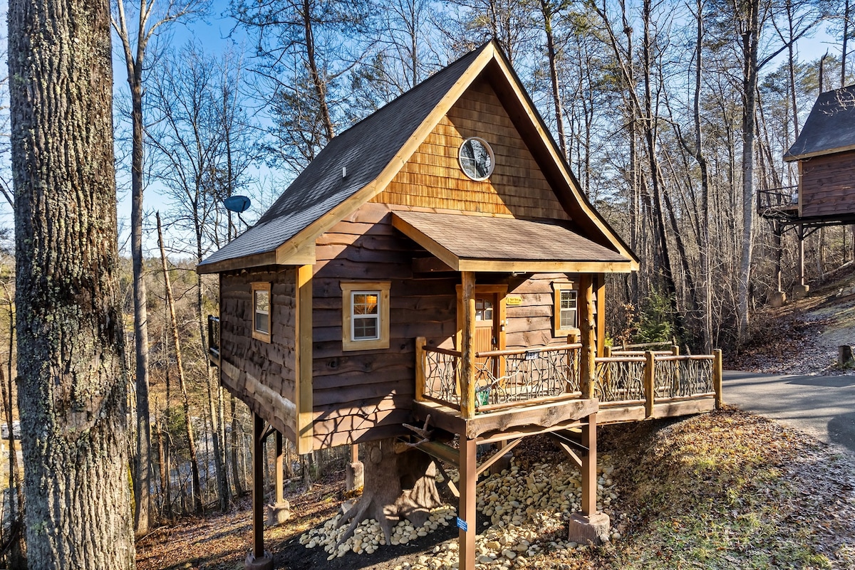 The exterior of the treehouse showcases a rustic wooden design, featuring a pitched roof and a large round window. A spacious porch extends from the front, supported by sturdy wooden beams. Surrounding trees provide a natural backdrop, enhancing the sense of seclusion.