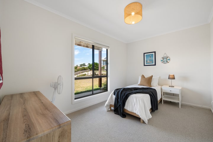 Კარგად განათებული საძინებელი ერთადგილიანი საწოლით ნეიტრალური ტონებით, დახვეწილი კულონის განათებით და დიდი ფანჯრით.