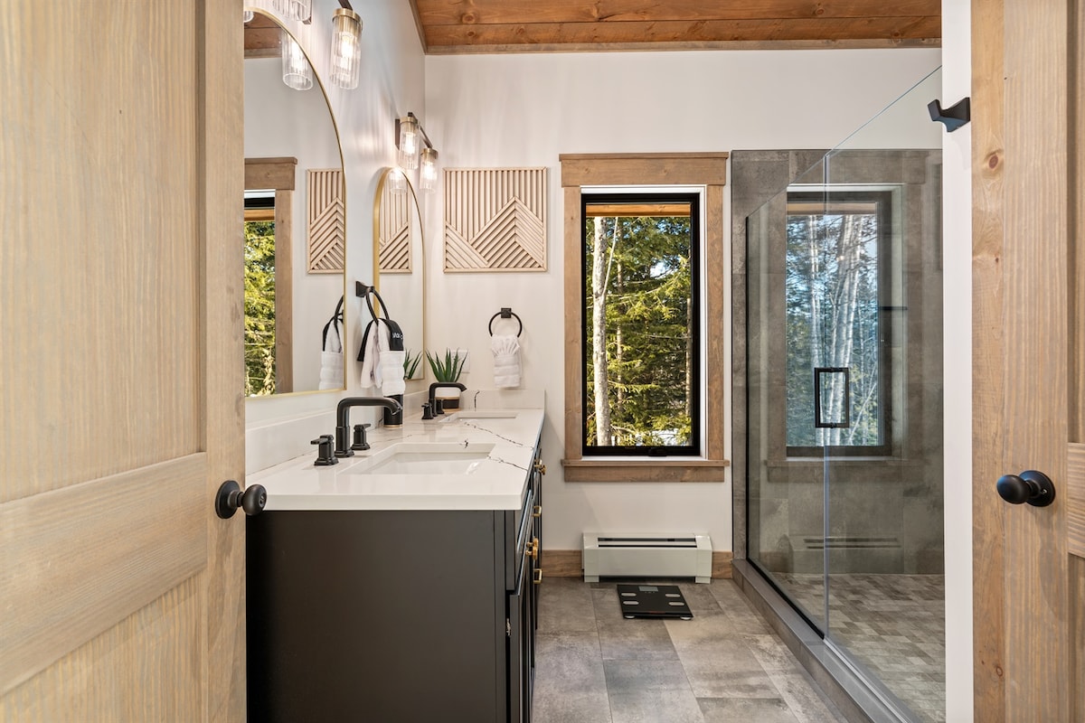 A modern bathroom features dual sinks set on a dark vanity, with stylish black fixtures. Large windows allow natural light and outdoor views into the space. A spacious walk-in shower with a clear glass door complements the sleek tile flooring and warm wood accents.