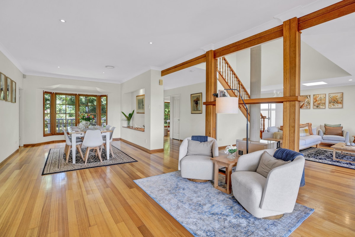 An inviting open living area features elegant hardwood flooring and natural light flooding through large windows. Comfortable armchairs are arranged on a textured rug, while a dining table is visible in the background, surrounded by chairs and decorated with potted greenery.