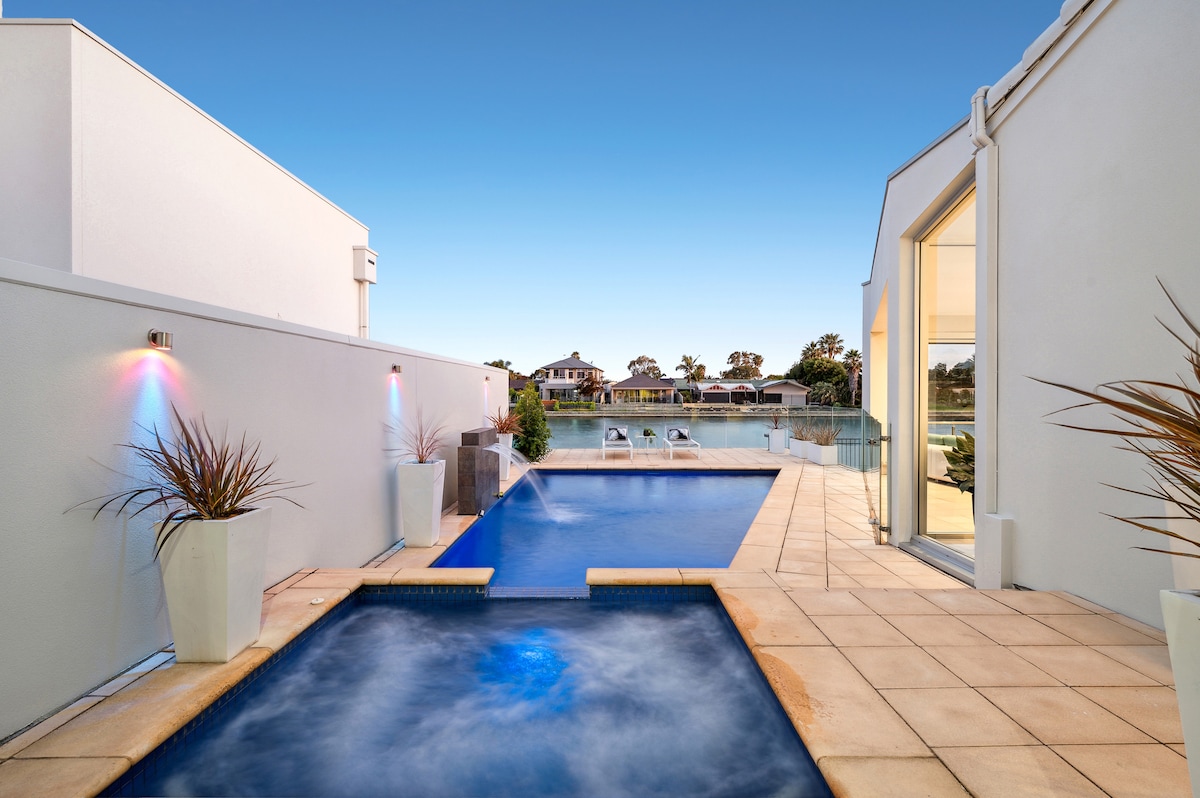 An outdoor area encompasses a solar-heated pool and spa, surrounded by spacious tiled terraces. Water features create a serene atmosphere, while modern planters add touches of greenery. A clear view of the lake and surrounding residences is visible in the background.