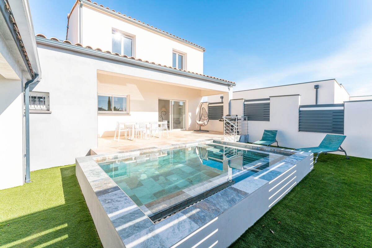 An outdoor area features a private pool surrounded by a stone patio. A set of loungers is positioned nearby, and a dining table with chairs is located under the covered terrace. Well-maintained grass borders the space, complemented by a clear blue sky.