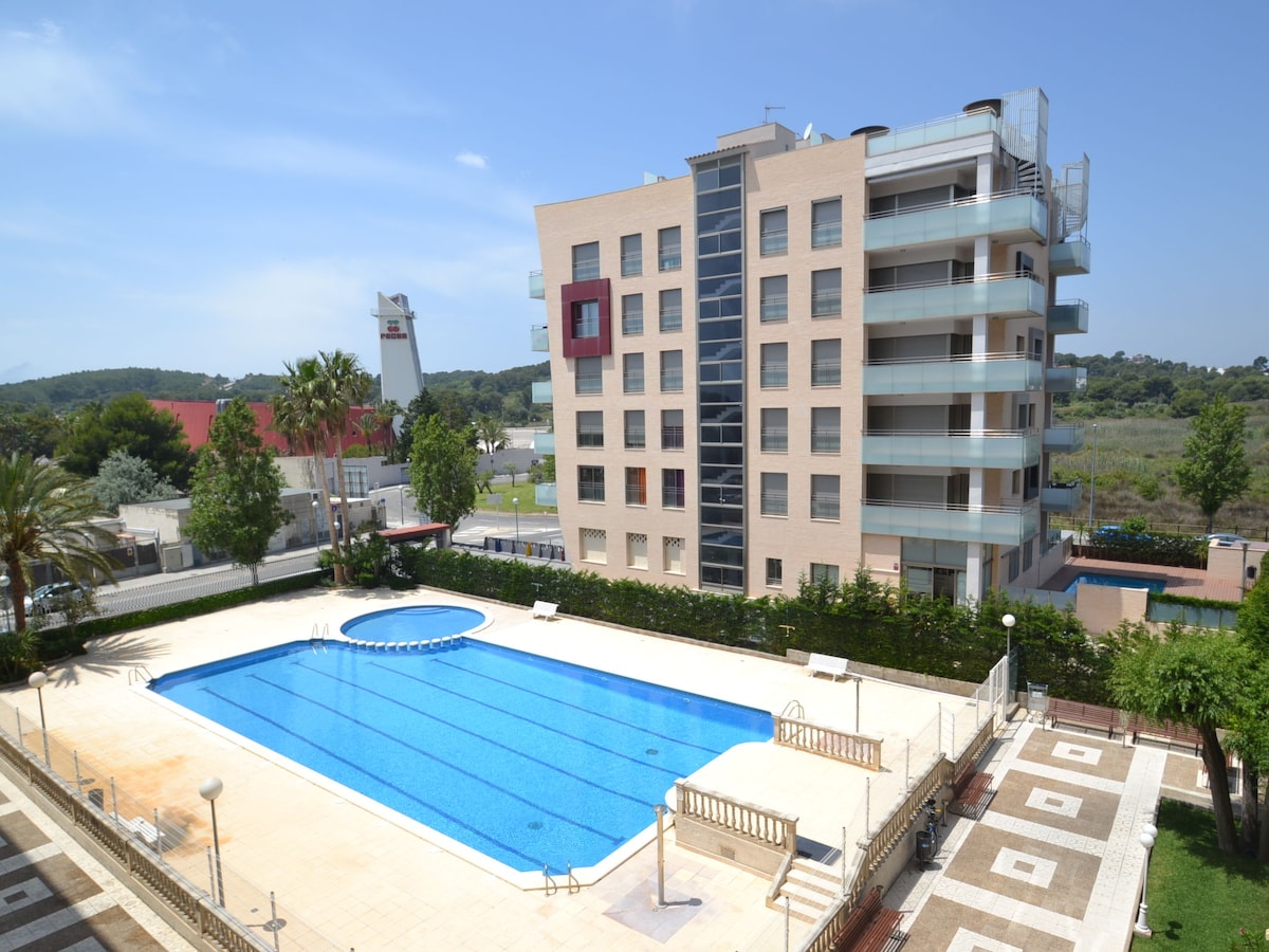A view of the swimming pool area of the Turquesa apartment block is showcased. The pool is rectangular with a circular section and is surrounded by a spacious deck. The modern building features multiple balconies and is set amidst greenery, providing a serene atmosphere.