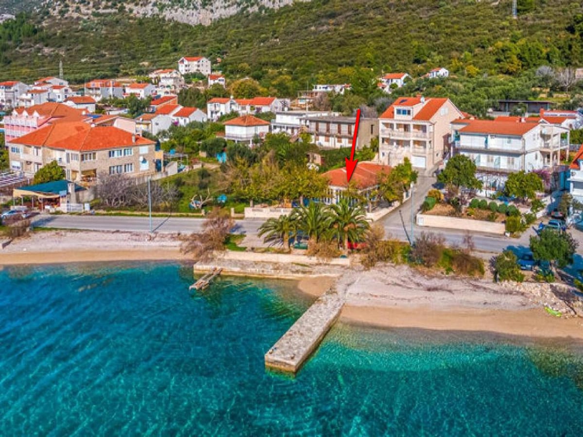 This aerial view captures the villa's location along the waterfront in Kuciste, with its red-roofed buildings visible amidst greenery. A sandy beach and a stone pier extend into the clear blue water, bordered by palm trees, enhancing the surrounding natural beauty.