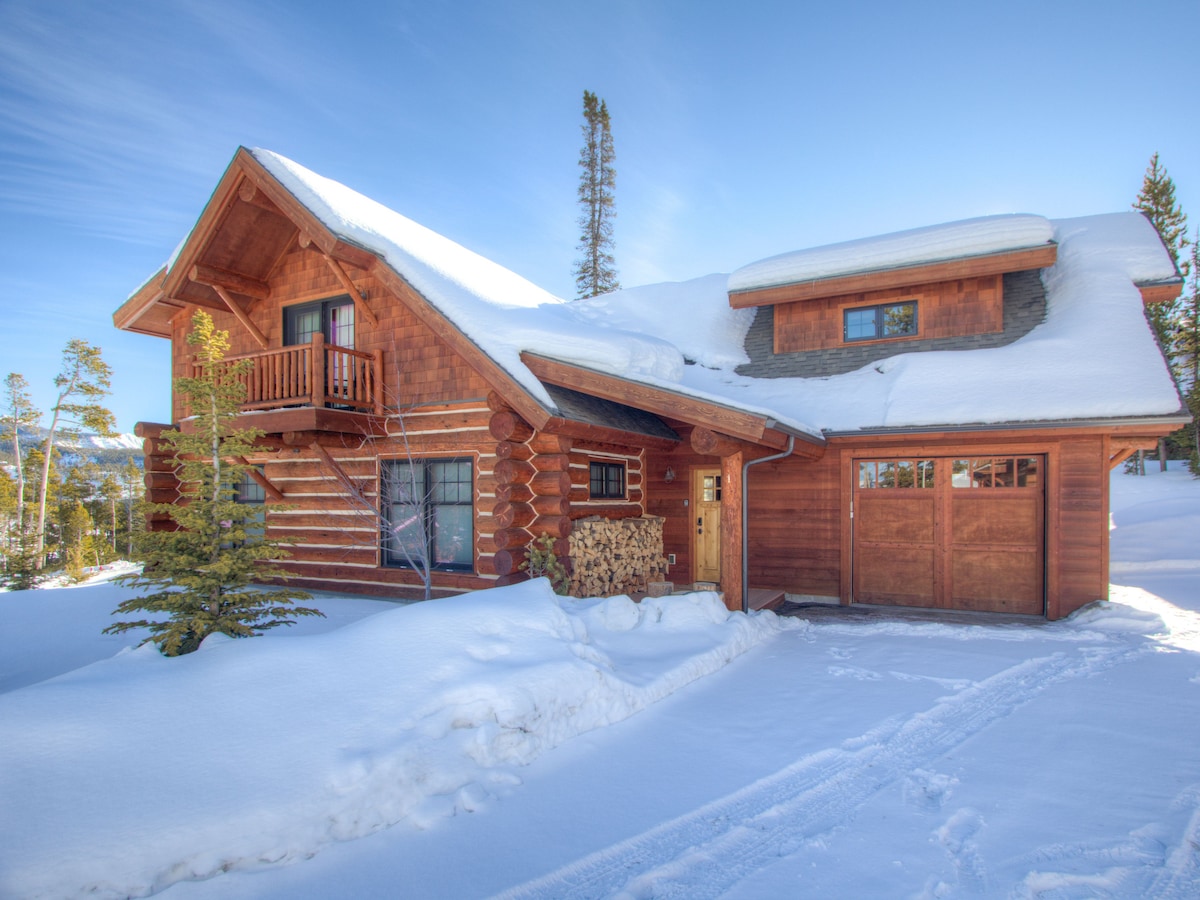 A spacious log cabin is nestled in a serene snowy landscape. The upper level features a covered balcony, while the main entrance is flanked by stacks of firewood. A single car garage is visible, contributing to the cozy mountain retreat ambiance.