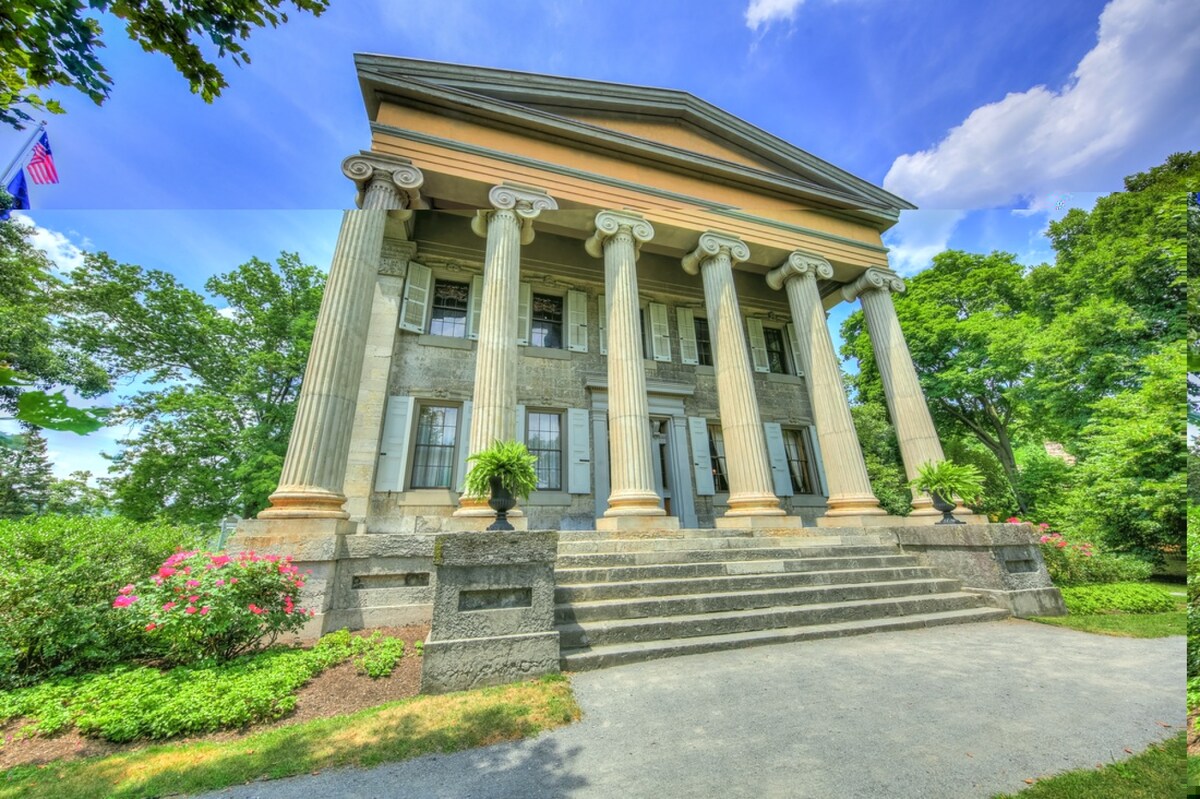 A grand building features large stone columns, supporting a classical portico. Lush greenery surrounds the structure, with vibrant flowering plants positioned at the base. The pathway leading to the entrance is framed by manicured gardens, providing an inviting approach.