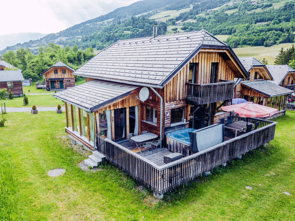 The exterior of the house features a wooden facade, complemented by a spacious terrace with a hot tub and outdoor seating. Surrounding grassy areas and a mountainous backdrop enhance the tranquil setting, while nearby structures are visible, contributing to a communal atmosphere.