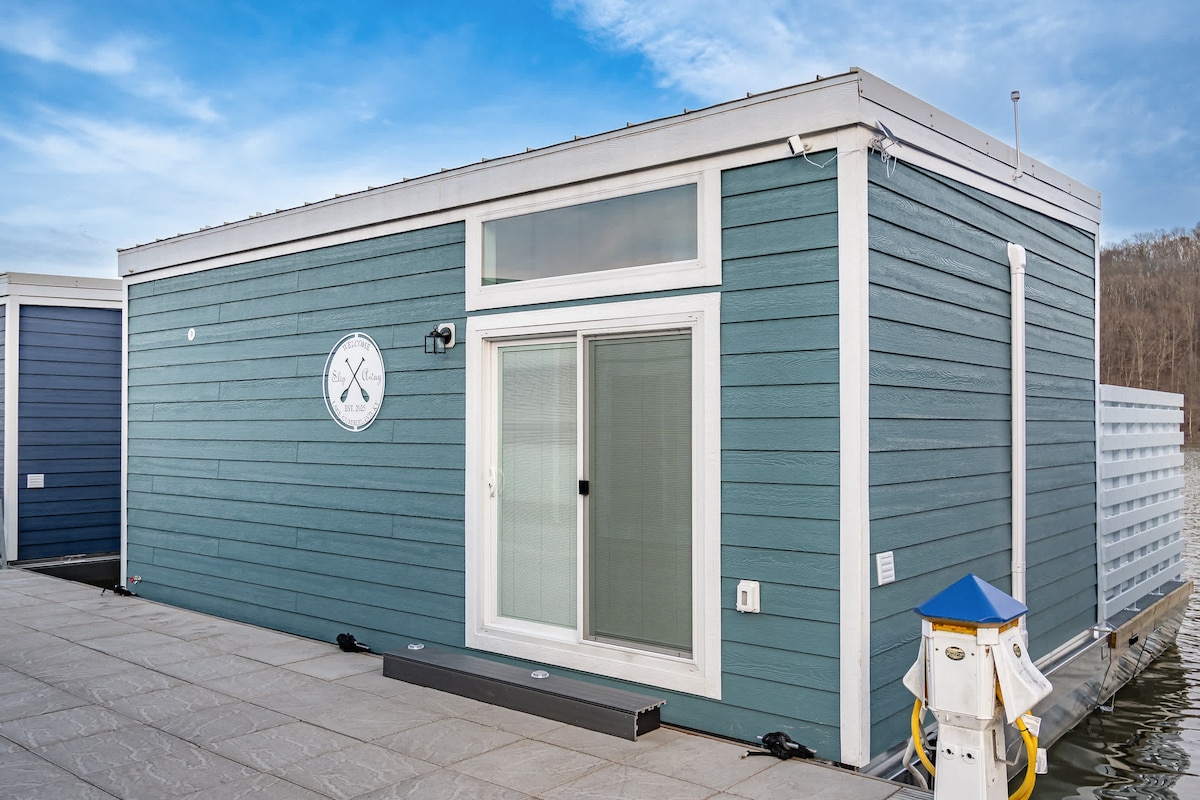A modern floating house is shown from the exterior, featuring a light blue façade with vertical siding. Large glass doors provide access to the interior, with a nautical-themed emblem displayed above. A walkway leads to the structure, alongside a docked boat mooring.