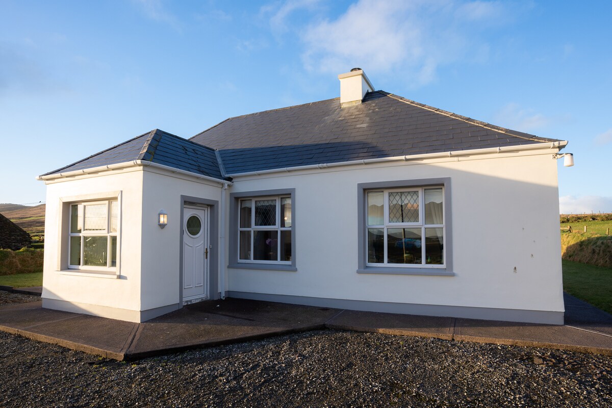 The exterior of a single-story cottage is displayed, featuring a gray slate roof and white walls. Large windows allow natural light to brighten the interior. The entrance is marked by a round door with decorative glass elements, leading to a welcoming atmosphere.