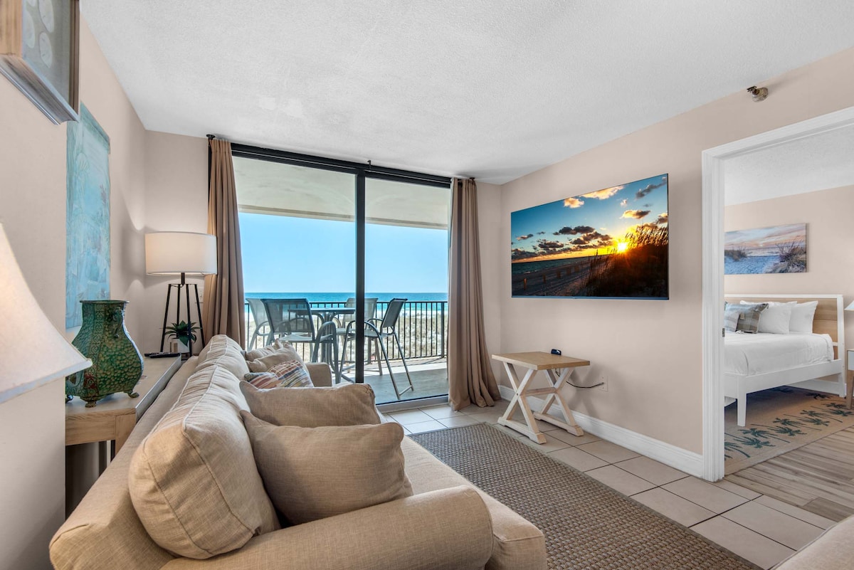 A bright living area is featured, showcasing a comfortable sofa and a flat-screen TV mounted on the wall. Large glass doors provide a view of the balcony overlooking the beach. Natural light illuminates the room, enhancing the neutral color palette.