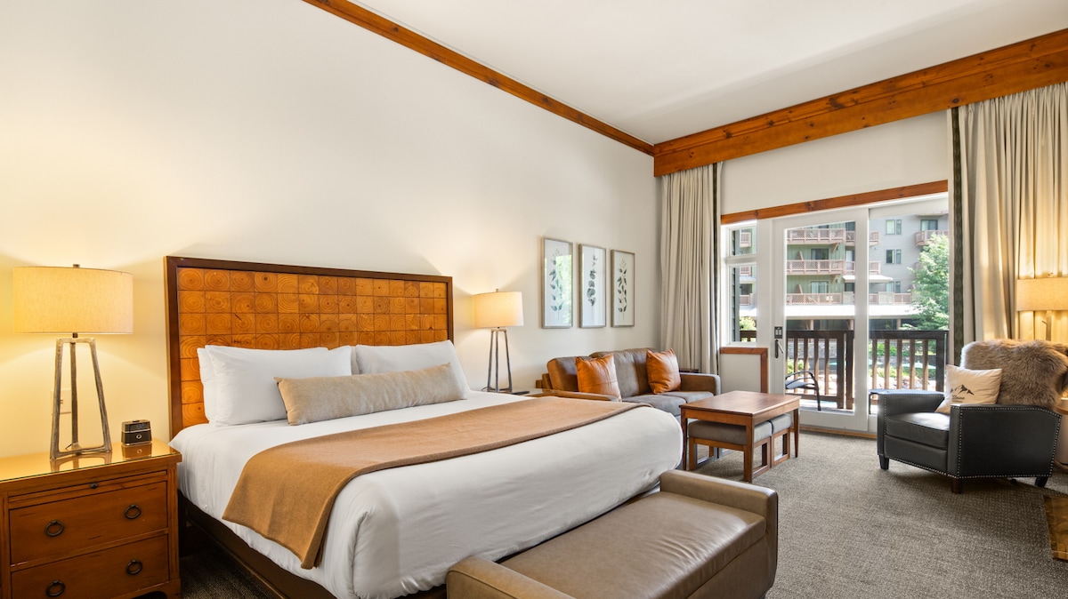 A spacious studio layout showcases a king-sized bed with neutral bedding, complemented by a queen sleeper sofa. Natural light fills the room through sliding glass doors that lead to a balcony. Seating arrangements include a comfortable armchair, a coffee table, and nightstand lamps.