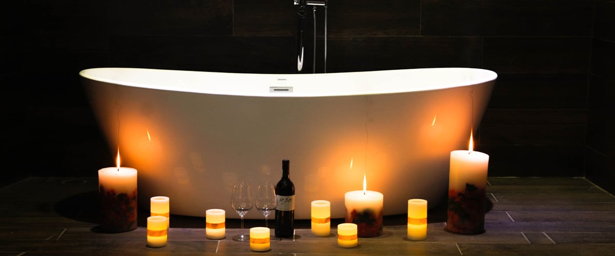 A freestanding white bathtub is positioned elegantly in the bathroom, surrounded by softly glowing candles in various sizes. A bottle of red wine and two wine glasses are placed nearby, enhancing the serene atmosphere created by the warm candlelight.