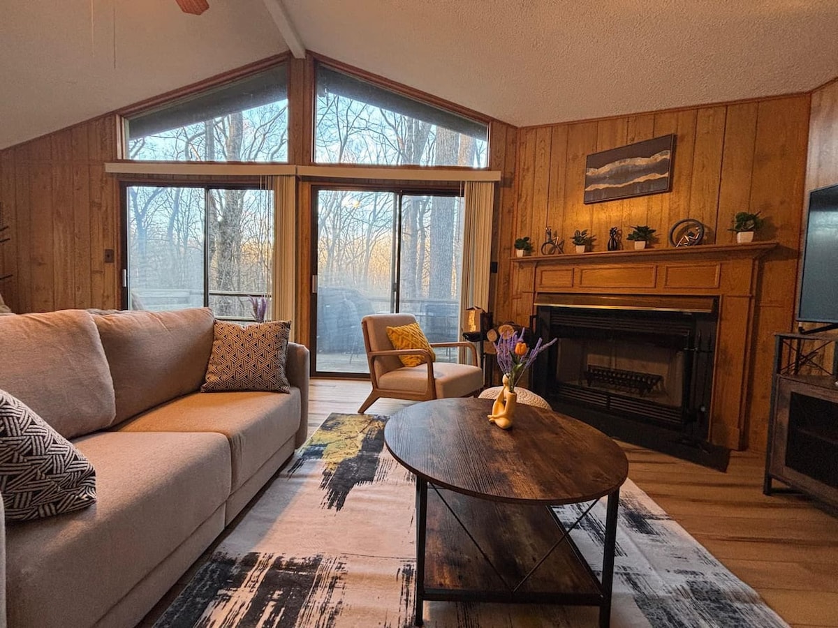 A cozy living area is adorned with wood paneling, showcasing large windows that frame forest views. A plush sofa, accented with throw pillows, is positioned beside a round coffee table. A fireplace is set against the wall, enhancing the inviting ambiance.